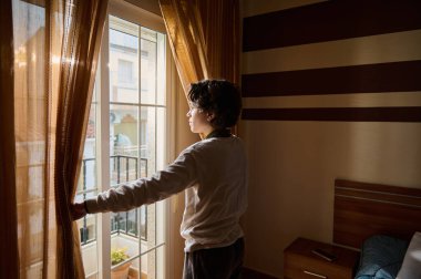 Bir kişi balkon penceresinin yanında durup, sabah güneşini içeri almak için perdeleri indiriyor. Çizgili duvarları, ahşap mobilyaları ve komodinin üzerinde bir telefonu olan rahat bir yatak odası..