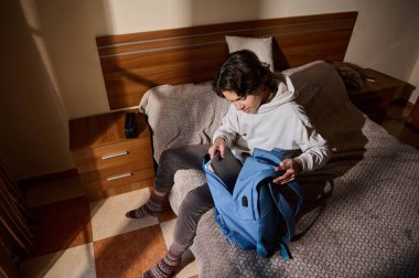 Genç bir adam sıcak, rahat bir yatak odasında bir yatakta oturur, mavi bir sırt çantası toplar ve defteri kontrol eder, çalışma, seyahat hazırlığı ve günlük rutin anları yakalar..