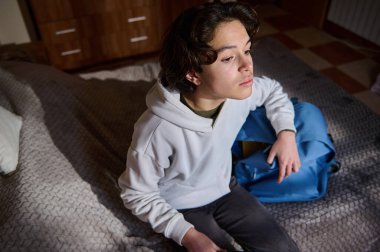 Beyaz kapüşonlu genç bir adam dokulu bir yatakta oturur, düşünceli bir şekilde yan tarafa bakar. Yakınlarda rahat bir yatak odasında mavi bir sırt çantası duruyor..