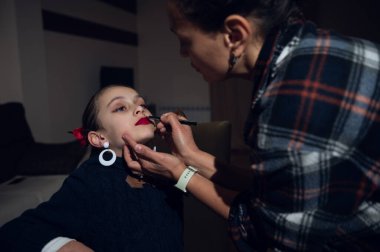 Bir makyaj sanatçısı, rahat bir stüdyoda oturan bir kadına, sahne arkası hazırlık anında güzellik, moda ve kendine güveni yakalayan koyu kırmızı ruj sürer..