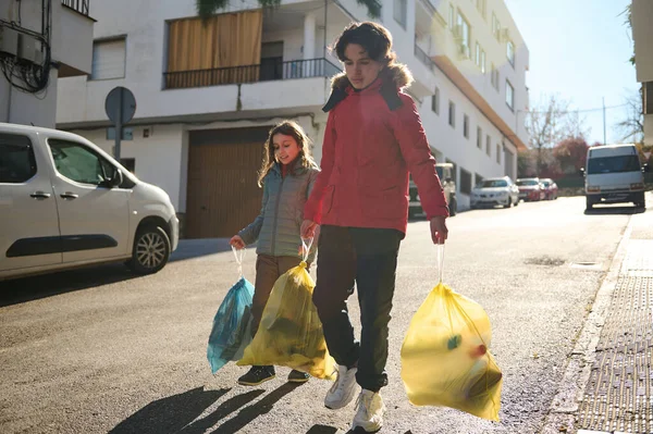 Ceketli iki kız güneşli bir şehir caddesinde yürüyor, her biri içinde eşyalarla dolu renkli bir çanta taşıyor, şehirde sıradan bir aile anı..