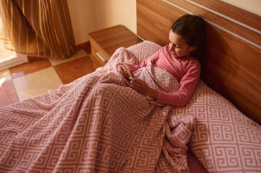 Bir kız yatakta, pembe bir geometrik battaniyeyle kaplanmış, rahat bir yatak odasında ahşap bir yatak başlığı ve sıcak bir ışık kullanarak gülümsüyor..