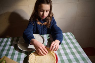 Mavi kazaklı genç bir kız, hamuru damalı bir masanın yanındaki kırmızı tabakta düzleştiriyor, sıcak, aile dostu bir pişirme anını yakalıyor sıcak bir ev mutfağında..