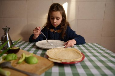 Genç bir kız sıcak bir mutfak masasında oturuyor, tabağında avokado olan gözlemeler yiyor. Sıcak ışık kahvaltı sahnesini dolduruyor..