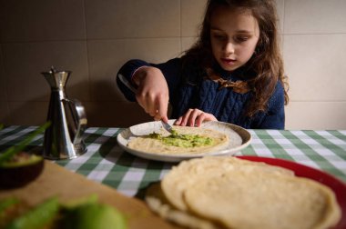Genç bir kız sıcak bir ekmeğin üzerine avokado sosu sürüyor. Sıcak bir aile mutfak masasında. Basit bir ev yemeği anının tadını çıkarıyor..