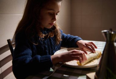 Genç bir kız sıcak bir mutfak masasında hamuru bir tabağa sarar, tortilla hazırlar ya da sarar..