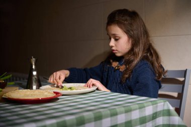 Genç bir kız sıcak bir evde yemek masasında oturuyor, tabaktan salata yiyor. Yeşil kareli masa örtüsü ve mavi kazak sıcak, samimi bir aile yemeği zamanı yaratır..
