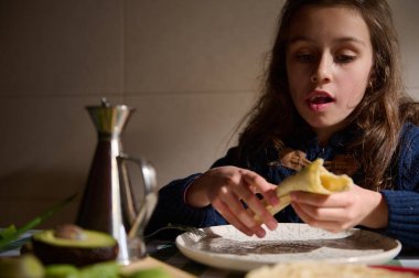 Genç bir kız sıcak bir mutfakta masada oturuyor, pastayı parçalıyor. Metal bir kahve demliği ve taze malzemeler sıcak bir aile kahvaltısı anını işaret ediyor..