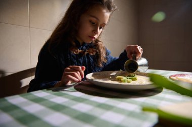 Koyu renk kazaklı genç bir kız sıcak bir mutfaktaki kareli bir masada avokado parçalarıyla bir tabağa sos döküyor..