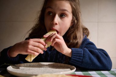 Mavi kazaklı genç bir kız sıcak bir mutfaktaki bir masada tortilla sargısı yer, ve sıradan bir yemek anını yakalar..
