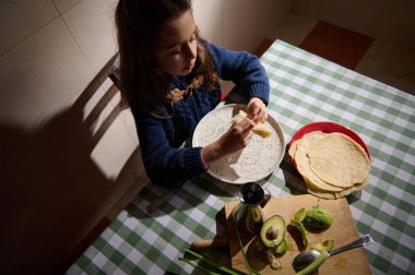 Genç bir kız damalı bir masada oturmuş krep, avokado, tortilla ve ekmekle basit bir kahvaltı ederken kahve fincanı da rahat bir sabah sahnesini tamamlıyor..