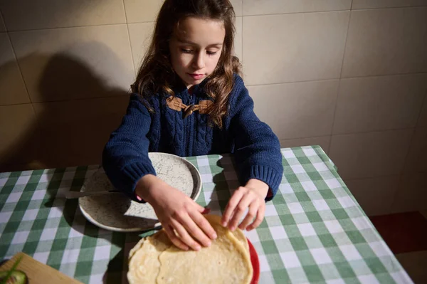 Mavi kazaklı genç bir kız, hamuru damalı bir masanın yanındaki kırmızı tabakta düzleştiriyor, sıcak, aile dostu bir pişirme anını yakalıyor sıcak bir ev mutfağında..