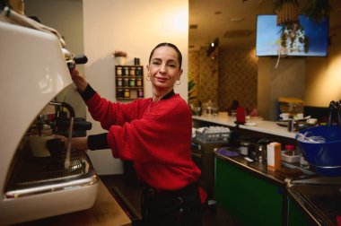Kırmızı kazaklı arkadaş canlısı bir kafe kahvecisi sıcak bir kafede müşterilere servis yapan bir espresso makinesi kullanıyor. Parlak tezgahlar, kahve aletleri ve yoğun, rahat bir atmosfer..