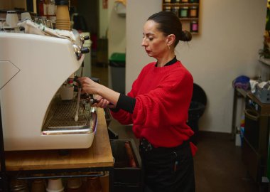 Kırmızı kazaklı İspanyol bir orta yaşlı barista sıcak bir kafede espresso makinesi kullanıyor. Yetenekli kahve yapıyor ve müşterilere dostça bir misafirperverlik sunuyor..