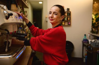 Kırmızı kazaklı bir İspanyol kafe sahibi tezgahın arkasında bir espresso makinesi çalıştırıyor. Sıcak, samimi bir kahve dükkanında kahve servisi yaparken içten bir gülümsemeyle gülümsüyor.
