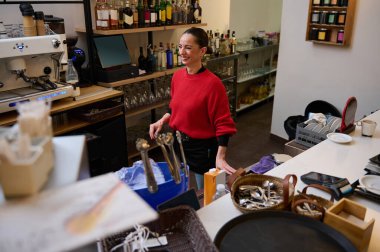 Kırmızı kazaklı İspanyol kafe sahibi kalabalık bir barın arkasında duruyor, rahat bir kafe ortamında içki ve bulaşıkları yönetirken gülümsüyor..