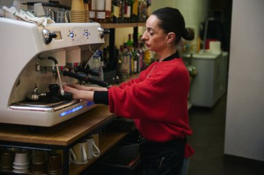 Kırmızı kazaklı bir İspanyol kafe sahibi barın arkasında büyük bir espresso makinesi işletiyor. Sıcak, modern bir kafede içki hazırlıyor..