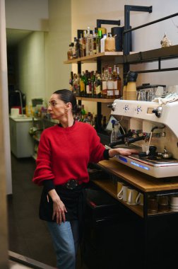 Parlak kırmızı kazaklı bir İspanyol kafe sahibi şişelerle çevrili, modern bir espresso makinesinin yanında duruyor. Odaklanmış görünüyor, hareketli bir kafede hizmet etmeye hazır..