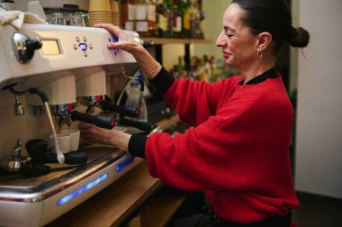 Kırmızı kazaklı bir kafe sahibi tezgahın arkasında profesyonel bir espresso makinesi işletiyor. Konukseverlik, küçük iş hayatı ve günlük kafe aktiviteleri sergiliyor..