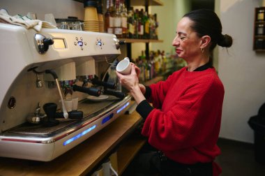 Kırmızı kazaklı orta yaşlı İspanyol bir kafe sahibi yoğun bir espresso makinesinde kahve yapıyor sıcak, misafirperverlik ve küçük bir aile kafeteryasının günlük telaşını iletiyor..