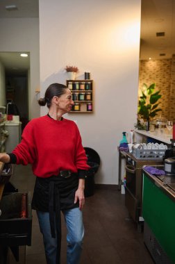 Kırmızı örgü süveter ve önlükle gülümseyen bir kafe sahibi, rafları, bitkileri ve tabakları olan sıcak ve yoğun bir kafede misafirleri karşılıyor..