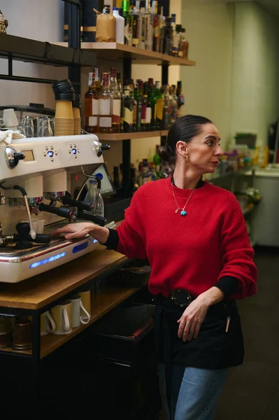Kırmızı kazaklı bir İspanyol kafe sahibi, rahat bir kafede bir espresso makinesinde çalışıyor. Arkasında raflar dolusu şişeler var. Konukseverlik ve küçük iş ortamı yansıtıyor..