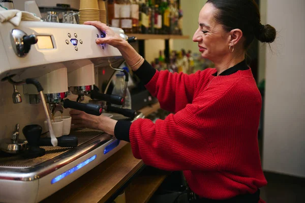 Kırmızı kazaklı bir kafe sahibi tezgahın arkasında profesyonel bir espresso makinesi işletiyor. Konukseverlik, küçük iş hayatı ve günlük kafe aktiviteleri sergiliyor..