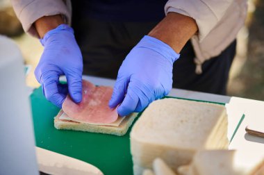 Eldivenli ellerin açık havada jambonlu sandviçi birleştirip yeşil bir kesme tahtasının üzerine jambon koyduğu yakın plan. Taze malzemeler ve günlük yemek hazırlama. Yemek hazırlama anı..