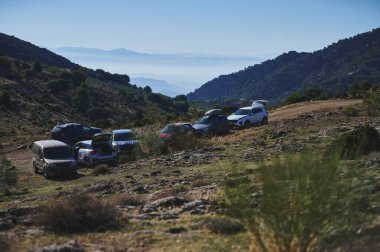 Uzaktaki dağlar sisli bir vadiye dönüşürken kayalık bir yamaçta bir sıra araç duruyor. Seyahat, macera ve doğal manzaralı açık hava eğlencesi için idealdir..
