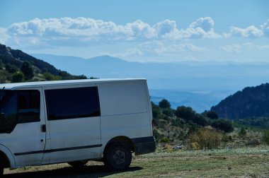 Kirli bir yol kenarına park edilmiş beyaz kargo minibüsü katmanlı dağlara ve mavi gökyüzüne karşı kurulmuş. Sakin, maceralı bir açık hava sahnesi seyahat ve keşif için mükemmel..