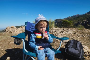 Küçük bir çocuk kayalık bir dağın tepesinde mavi bir kamp koltuğunda oturur ve içkisini yudumlar. Güneşli gökyüzünün altında engebeli bir arazide bir sırt çantası duruyor..