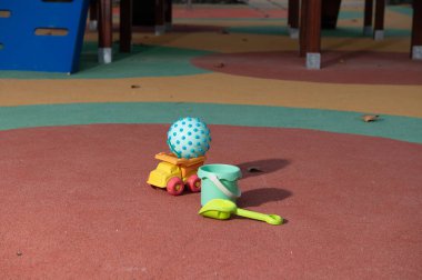 Renkli kum oyuncakları, kauçuk bir oyun alanı yüzeyinde dinleniyor. Noktalı bir top, parlak bir kova, kürek ve kamyon eğlenceli açık hava anlarını çağrıştırıyor..
