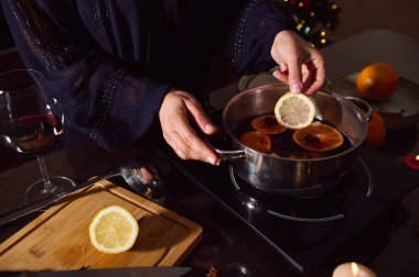 Sıcak, şenlikli bir an. Koyu renkli, zarif bir elbise giymiş bir kadın ocakta narenciye dilimleri, mum ışığı ve yakınlarda bir bardak şarap hazırlıyor..