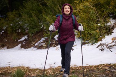 Kestane rengi kapüşonlu ceketli bir kadın karlı bir ormanda yürüyüş yapıyor, yürüyüş direkleri kullanıyor ve sırt çantası taşıyor. Gülümsüyor, soğuk hava macerasının tadını çıkarıyor..