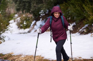 Bir kadın ormanda karla kaplı bir patikada yürüyüş yapar, yürüyüş direkleri kullanır ve kahverengi bir ceket ve sırt çantası giyer. Açık hava macerası ve temiz hava kış ortamında..