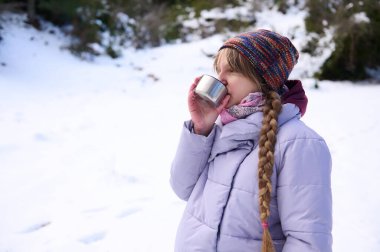 Mor kabarık ceketli genç bir kız, karlı bir arazide, örgülü saçları ve örgü şapkasıyla metal bir bardağı yudumluyor. Açık havada huzurlu bir kış anı.
