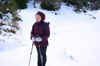 Koyu renk ceketli bir kadın, kar kaplı bir patikada yürüyüş direkleri ile kış yürüyüşüne hazırdır. Sırt çantası taşıyor, tek başına geziyor ve soğuk hava macerası yaşıyor..