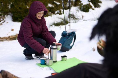 Sıcak ceketli bir kadın karda diz çöker, termosla kahve hazırlar ve bir sırt çantasının yanında fincanlar ve karlı bir günde yürüyüş direkleri. Barışçıl kış macerası.