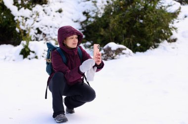 Kestane rengi ceketli ve sırt çantalı bir kadın, karlı bir arazide çömeliyor, elinde bir selfie çekmek için akıllı telefon tutuyor. Eldivenleri tak, ekrana odaklan.