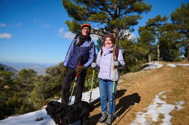 İki yürüyüşçü, siyah bir köpekle karlı bir dağ yolunda yürüyor, güneşli havanın ve geniş manzaranın tadını çıkarıyorlar..