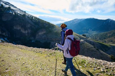 Sırt çantalı iki yürüyüşçü, parlak mavi bir gökyüzünün altında yükselen karlı zirveler gibi, ellerinde yürüyüş direkleriyle kayalık bir dağ yolunda yürür..