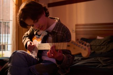 Genç bir adam güneşli bir yatak odasında, elektro gitar çalmaya odaklanmış bir şekilde bir yatakta oturuyor. Sıcak doğal ışık, günlük kıyafet, samimi ev reçeli..