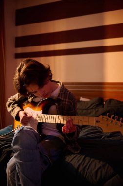 Genç bir müzisyen sıcak bir odada elektro gitar tıngırdatarak yatakta oturuyor. Çizgili duvar, müzik, hobi ve yaratıcı ilham için samimi bir ortam yaratır..