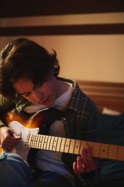 Ekose gömlekli genç bir adam sıcak, rahat bir ev ortamında elektro gitarını nazikçe tıngırdatıyor..