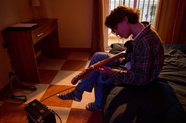 Ekose gömlekli genç bir adam sıcak, loş bir yatak odasında bir yatakta oturuyor ve yanında küçük bir amfiyle elektro gitar çalıyor..