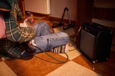 Kotla ve ekose gömlekle fayanslı zeminde oturan bir insan sıcak, rahat bir ev müziği köşesinde kompakt bir amplifikatöre bağlı elektro gitar çalar..