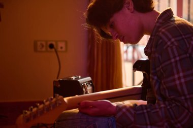 Genç bir adam sıcak bir ışıkta oturuyor, bir duvar prizi ve pencerenin yanında elektro gitar tıngırdatıyor..