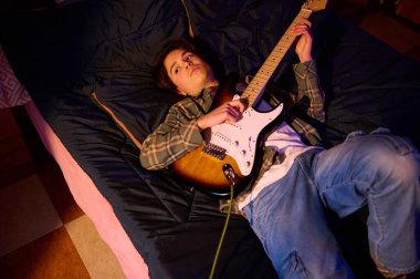 Genç bir adam, sıcak ve samimi bir ev ortamında, güneş patlaması yaşayan bir gitarla, bir yatakta dinleniyor ve müzikten zevk alıyor..