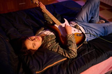 Ekose gömlekli ve kotlu genç bir adam bir yatağa uzanıyor, elleri elektro gitarda, sıcak, samimi bir evde..