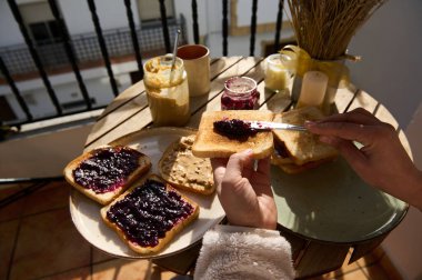 Fıstık ezmesi ve reçelli tost, reçel kavanozları ve kırsal dekor içeren parlak bir balkon kahvaltısı. Tost üzerine reçel serpme, ev yapımı konfor ve güneşli sabah enerjisini yakalama anı..